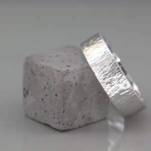 May include: Two silver wedding bands with a textured finish, one on top of the other, resting on a gray concrete block.