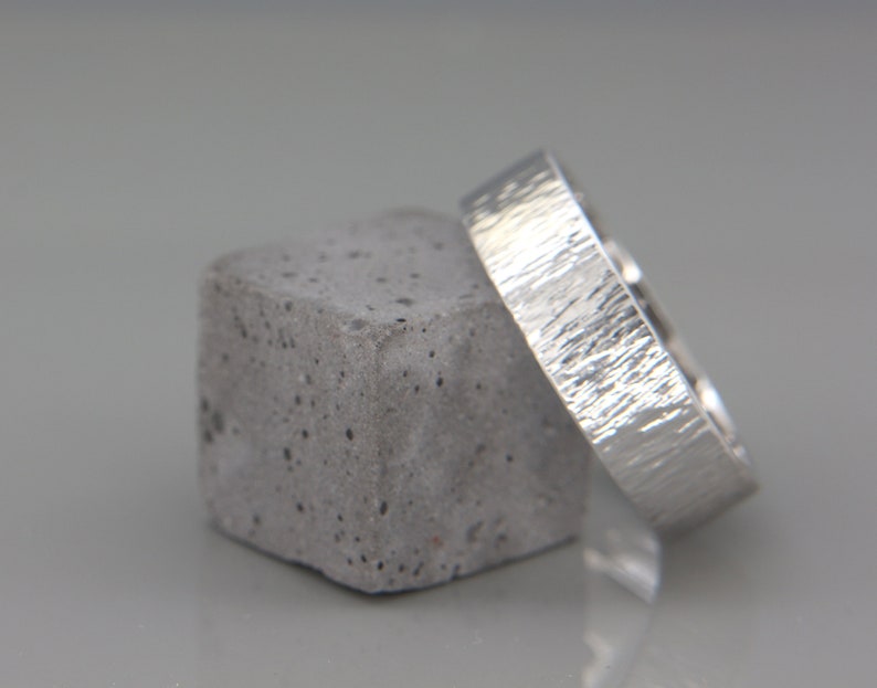 May include: Two silver wedding bands with a textured finish, one on top of the other, resting on a gray concrete block.