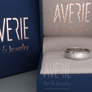 May include: A silver wedding band with a brushed finish and a beveled edge. The ring is in a blue box with the text "Averie Art & Jewelry" on it.