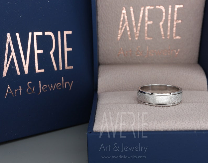 May include: A silver wedding band with a brushed finish and a beveled edge. The ring is in a blue box with the text "Averie Art & Jewelry" on it.