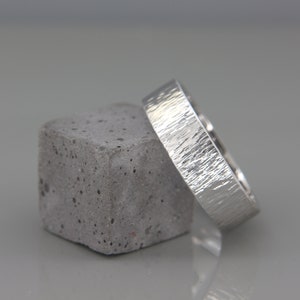 May include: A silver wedding band with a textured finish, resting on a gray concrete block.