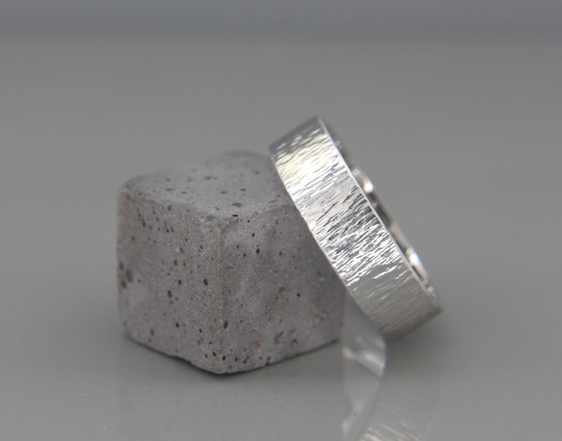 May include: A silver wedding band with a textured finish, resting on a gray concrete block.