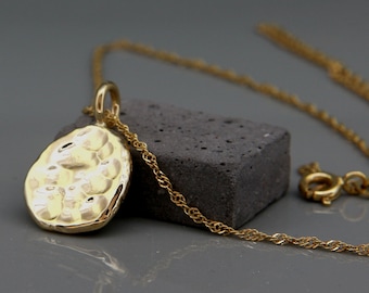 May include: A gold necklace with a hammered, oval-shaped pendant. The chain is a delicate, rope style.