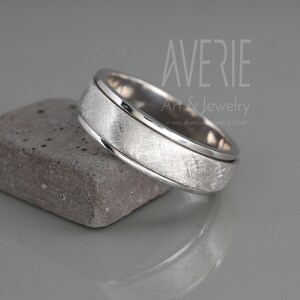 May include: A silver wedding band with a brushed finish and a polished edge. The band is on a gray textured surface.
