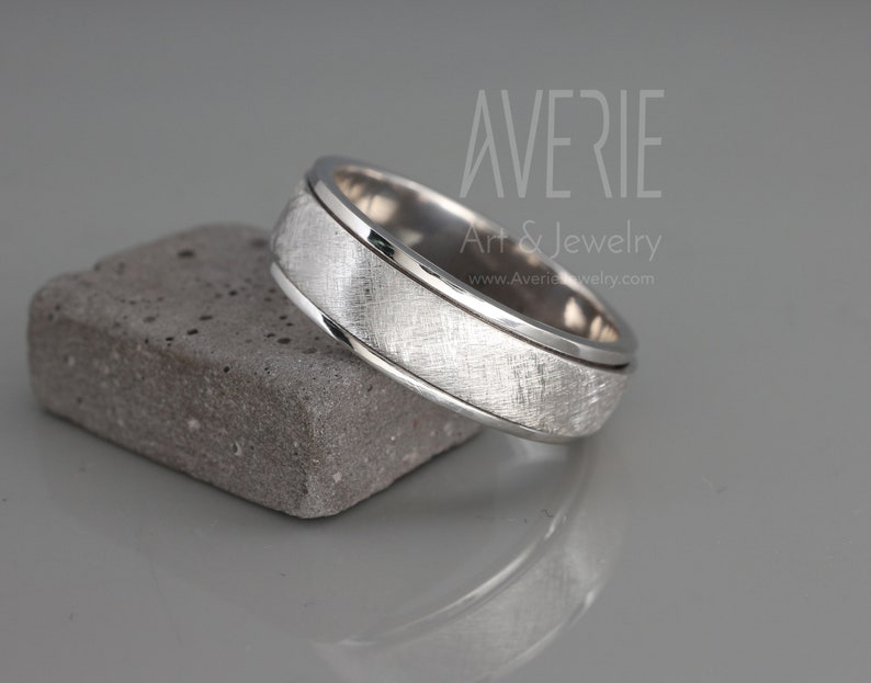 May include: A silver wedding band with a brushed finish and a polished edge. The band is on a gray textured surface.