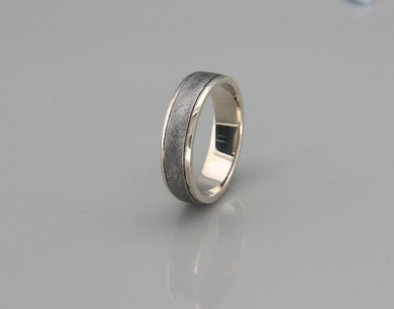 May include: A silver wedding band with a textured, dark gray finish. The band has a polished edge.