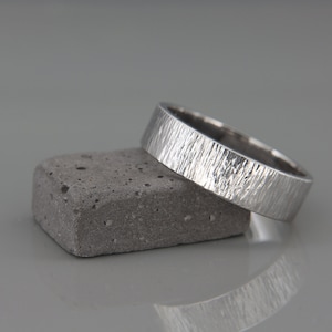 May include: A silver wedding band with a textured finish, resting on a gray concrete block.