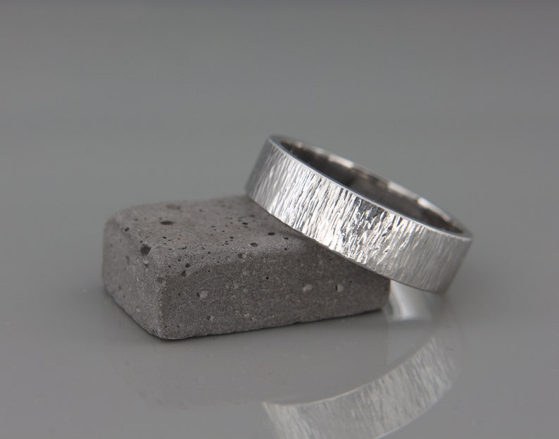 May include: A silver wedding band with a textured finish, resting on a gray concrete block.