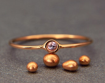 May include: A delicate gold ring with a small pink gemstone set in the center. The ring is resting on four small, round, gold beads.