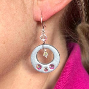 May include: Silver-toned hoop earrings with three pink crystals and a dangling crystal accent. The earrings have a circular design with a small crystal at the top and are worn on the ear.