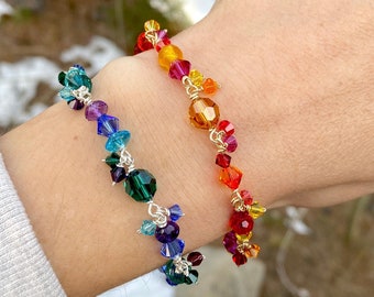 May include: Two beaded bracelets, one in a rainbow of colors and the other in shades of blue, purple, and green, are displayed on a wrist. The bracelets feature small, faceted beads strung together with silver-colored wire, creating a delicate and colorful accessory.