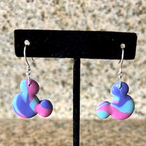 May include: A pair of dangle earrings in the shape of a cartoon mouse head. The earrings are made of a marbled polymer clay in shades of pink, blue, and purple. The earrings hang from silver-colored hooks.