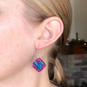 May include: A pair of square-shaped earrings with a pink glitter border and a blue glitter design. The earrings are hanging from silver hooks. The earrings are being worn on a person's ear.