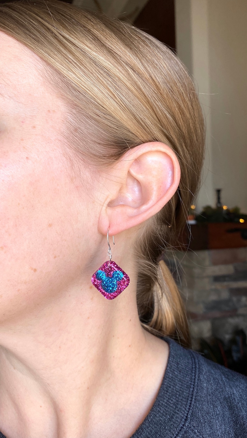 May include: A pair of square-shaped earrings with a pink glitter border and a blue glitter design. The earrings are hanging from silver hooks. The earrings are being worn on a person's ear.