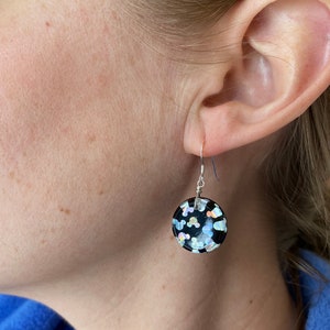 May include: A black, round earring with silver-tone wire and a hook. The earring is decorated with iridescent confetti-like shapes. The earring is shown on a person's ear.