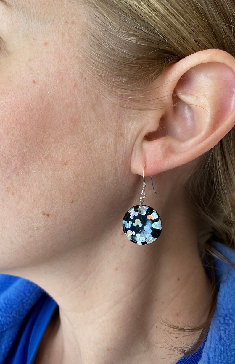May include: A black, round earring with silver-tone wire and a hook. The earring is decorated with iridescent confetti-like shapes. The earring is shown on a person's ear.