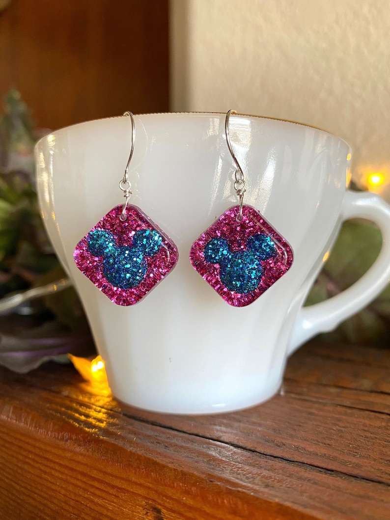 May include: A pair of dangle earrings featuring a pink glitter square with a blue glitter Mickey Mouse head design. The earrings have silver-colored hooks and are displayed in a white teacup.