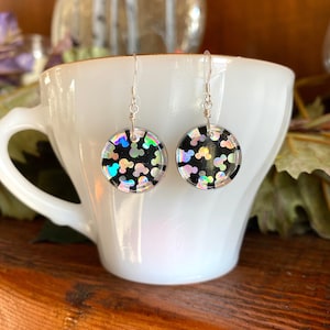 May include: A pair of round dangle earrings with a black background and iridescent Mickey Mouse shapes. The earrings are suspended from silver-colored fish hook ear wires. The earrings are displayed in a white teacup.