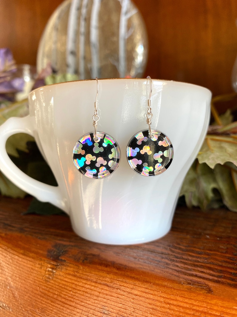 May include: A pair of round dangle earrings with a black background and iridescent Mickey Mouse shapes. The earrings are suspended from silver-colored fish hook ear wires. The earrings are displayed in a white teacup.