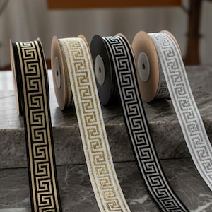 May include: Four reels of decorative ribbon featuring a Greek key pattern in gold, white, black, and silver. The ribbons are displayed on a grey marble surface, showcasing their intricate designs and colour variations.