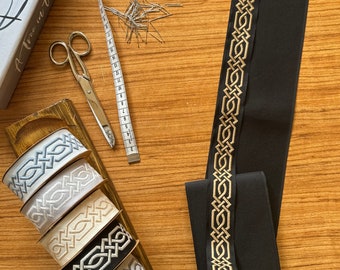 May include: Five spools of black, white, gray, and beige ribbon with a gold Celtic knot design. The ribbon is on a wooden surface with a measuring tape, scissors, and a jar of pins.