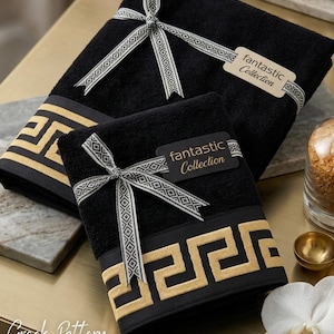 May include: A set of two black towels featuring a gold Greek key pattern border. Each towel is tied with a grey and white patterned ribbon and includes a tag labeled fantastic Collection. The towels are folded and stacked on a flat surface.