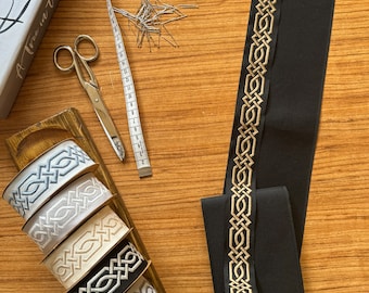 May include: Five spools of black, white, gray, and beige ribbon with a gold Celtic knot design. The ribbon is on a wooden surface with a measuring tape, scissors, and a jar of pins.