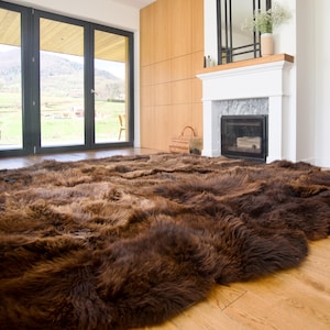 May include: A large, rectangular area rug made of thick, long-pile sheepskin in a deep, rich brown color. The rug features a natural, shaggy texture with varied tones of dark chocolate and mahogany brown, covering a light-colored wooden floor.