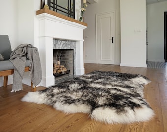 May include: A large, rectangular sheepskin rug featuring a long-pile texture with a variegated pattern of black, charcoal, and white fur. The rug has an irregular, natural edge shape and is positioned on a light brown wooden floor in a room.