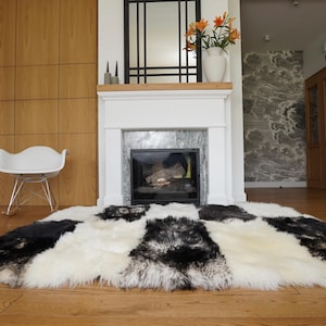 May include: A large, rectangular area rug with a patchwork design of black, white, and gray fur. The rug is placed in front of a white fireplace with a black framed mirror above it.