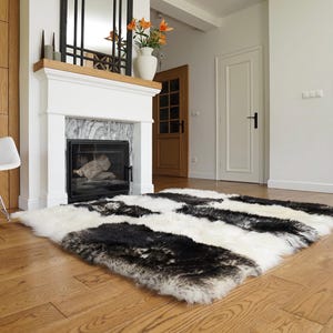 Puede incluir: Una alfombra rectangular de piel de oveja con un patrón en blanco y negro está sobre un suelo de madera. La alfombra está en un salón, cerca de una chimenea y una mecedora blanca. La alfombra mide aproximadamente 152 cm por 203 cm.