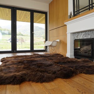 Puede incluir: Una alfombra de piel de oveja marrón con pelo largo y suave, colocada sobre un suelo de madera en una sala de estar moderna con chimenea y grandes ventanales.