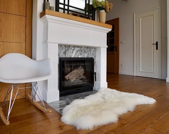 May include: A white rocking chair with a molded seat and chrome wire base with wooden rockers, positioned next to a white fireplace with a gray marble surround and a fluffy white sheepskin rug resting on the wooden floor in front of it.