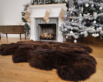 Op de afbeelding: Een bruin nep bontkleed voor een open haard met kousen die aan de schoorsteenmantel hangen. Op de achtergrond staat een kerstboom met witte kerstballen.