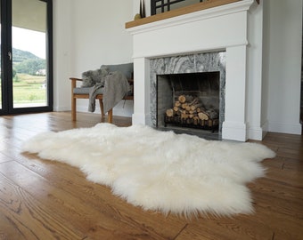 May include: A long-haired, off-white sheepskin rug with a natural, irregular shape and a thick, plush texture. The rug is positioned on a wooden floor, showcasing its dense, soft fibers and creamy white color throughout the entire surface area.