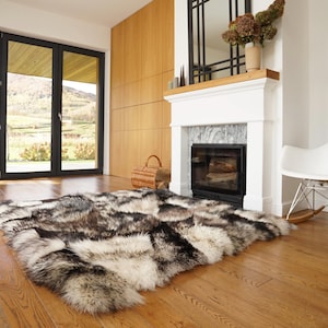 Peut inclure: Un grand tapis rectangulaire en fausse fourrure dans les tons de noir, gris et crème est posé sur un parquet. Le tapis est au centre d'un salon avec une cheminée, une chaise à bascule blanche et de grandes fenêtres donnant sur un paysage.