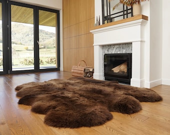 May include: A large, dark brown sheepskin rug with a thick, plush texture and an irregular, natural hide shape. The rug features dense, soft fur fibers and is laid flat on a light-toned wooden floor, showcasing its deep, uniform color.