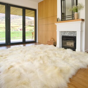 May include: A large, off-white sheepskin rug with long, thick, and soft fur fibers. The rug is spread across a light-colored wooden floor, featuring a natural, irregular shape with a dense, shaggy texture that covers a significant area.
