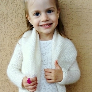 Può includere: Una bambina che indossa un cardigan bianco lavorato a maglia con un collo a scialle. Sta sorridendo e guardando la telecamera.