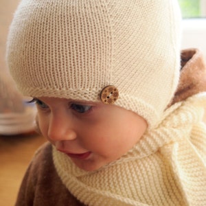 Peut inclure: Gros plan d'un bébé portant un bonnet et une écharpe en tricot blanc. Le bonnet est orné d'un bouton en bois avec un motif de flocon de neige.