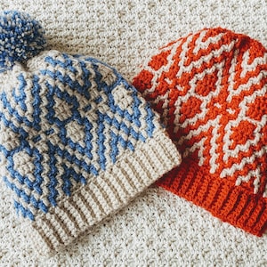 Puede incluir: Dos gorros de punto de ganchillo, uno con un patrón azul y beige y un pompón blanco, el otro con un patrón naranja y beige.