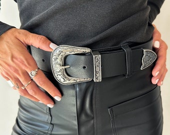 May include: A black leather belt with a silver, ornate buckle. The belt is worn over a black ribbed shirt and black pants.