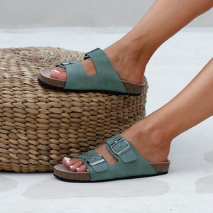 May include: A pair of sage green leather sandals with two adjustable buckles. The sandals have a cork footbed and are worn on a person's feet.