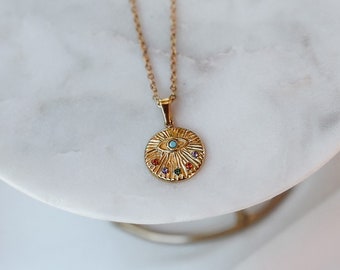 Puede incluir: Collar de oro con un colgante redondo con un diseño de ojo turco con piedras preciosas de colores. El colgante está sobre una superficie de mármol blanco.