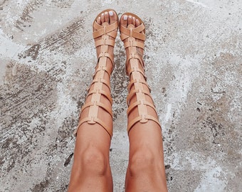 May include: A pair of tan leather gladiator sandals with straps up the leg. The sandals are worn by a person wearing a black skirt.