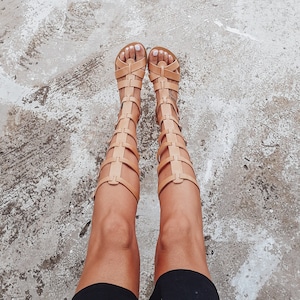 Może przedstawiać: Para brązowych skórzanych sandałów gladiatorów z paskami sięgającymi do nogi. Sandale nosi osoba w czarnej spódnicy.