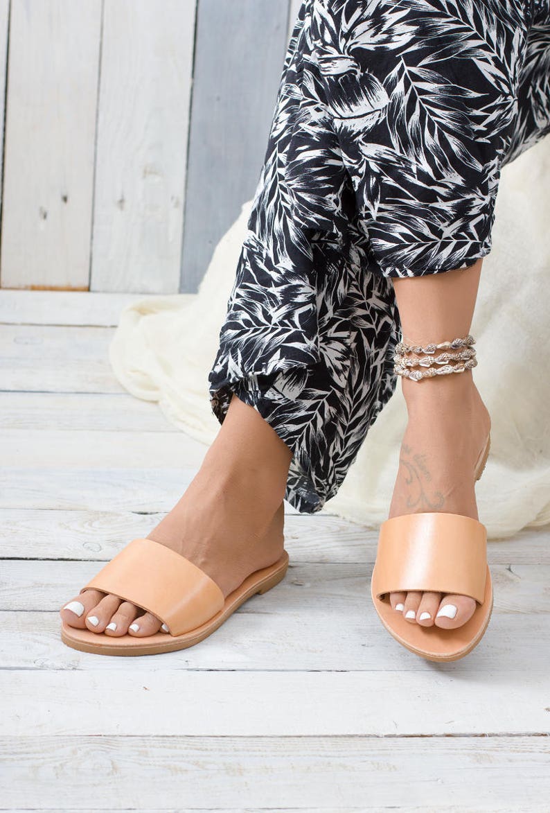 May include: A pair of light brown leather slide sandals with a simple design. The sandals are worn by a person with a black and white patterned pants.