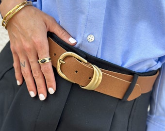 Peut inclure: Une ceinture en cuir marron avec une boucle dorée. La ceinture est portée sur un pantalon noir.