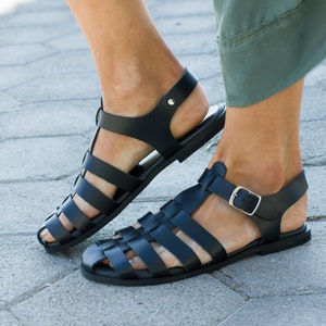 Puede incluir: Un par de sandalias de cuero negro con cierre de hebilla y dise&ntilde;o de correa tejida. Las sandalias se llevan en los pies de una persona.