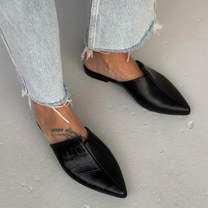 Può includere: Mules nere in pelle con punta. Un mule presenta un motivo strutturato, simile alla pelle di coccodrillo, sulla tomaia. Le scarpe sono indossate con jeans blu chiaro con orli sfilacciati. Lo sfondo è una superficie grigio chiaro.